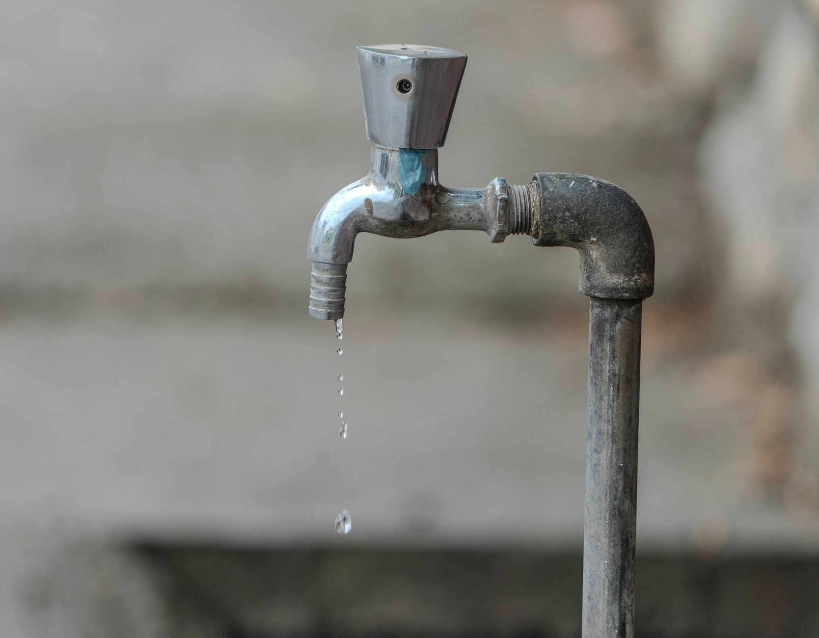 Robinet métallique au bout d’un tuyau vertical, avec quelques gouttes d’eau qui tombent devant un fond gris flou.
