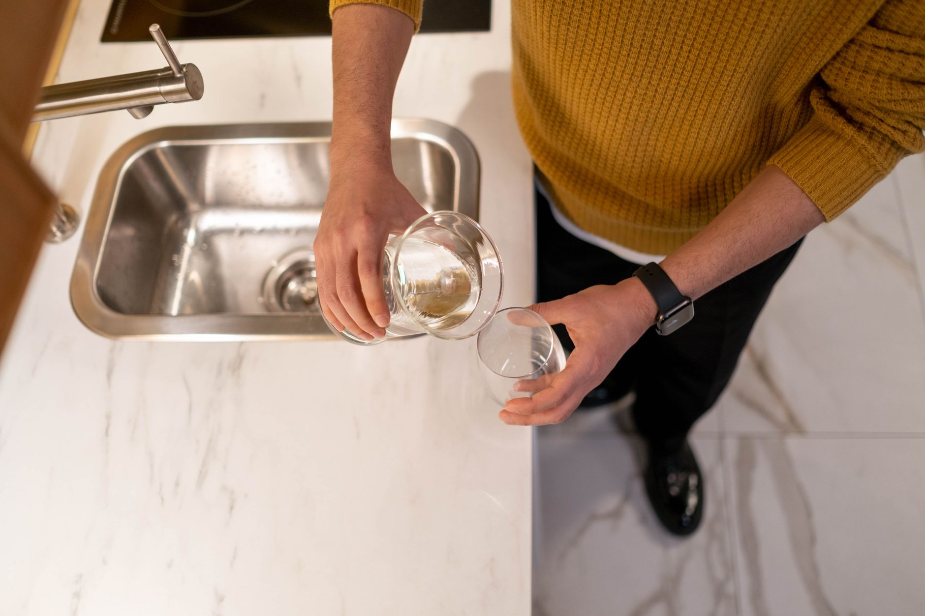 Vue de dessus d’une personne en pull jaune versant un liquide clair d’une carafe dans un verre, près d’un évier en inox.